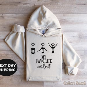 May include: A sand-colored hoodie with a black graphic of three wine openers and the text "My Favorite Workout".