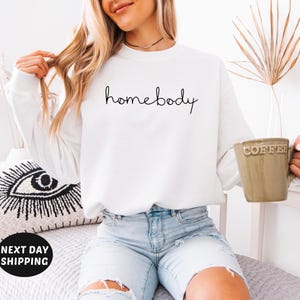 May include: White long-sleeved sweatshirt with the text "homebody" written in black cursive script.