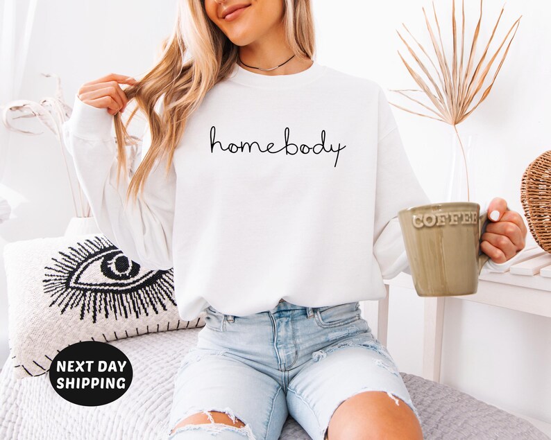 May include: White long-sleeved sweatshirt with the text "homebody" written in black cursive script.