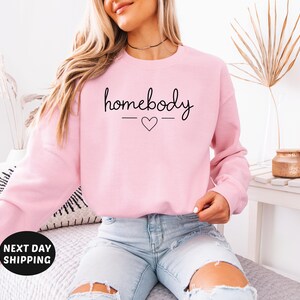 May include: A pink sweatshirt with the text "homebody" in black script with a small heart underneath.