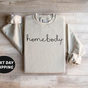 May include: A sand-colored sweatshirt with the text "homebody" written in black cursive.