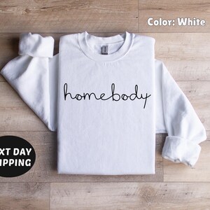 May include: White crew neck sweatshirt with the text "homebody" in black script.