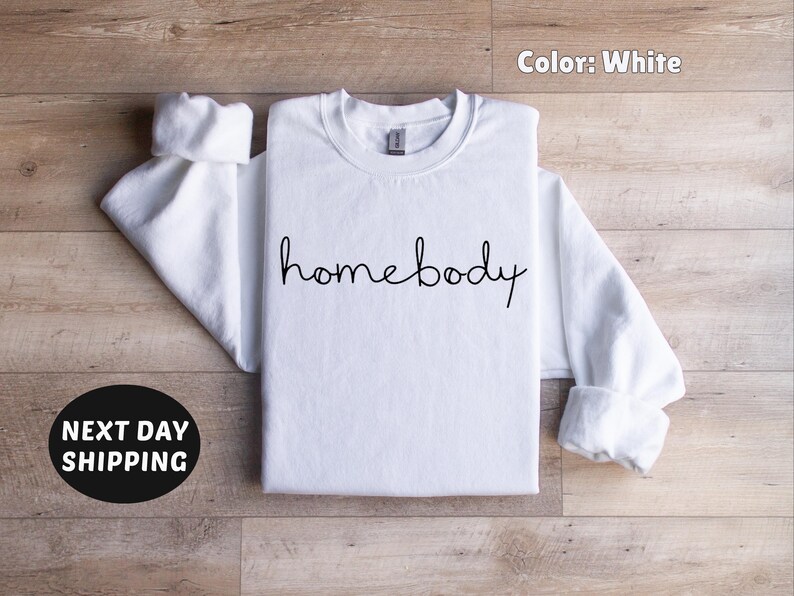May include: White crew neck sweatshirt with the text "homebody" in black script.