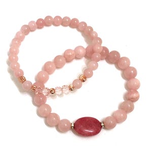 May include: Two pink rose quartz bracelets with gold accents. One bracelet has a larger, oval-shaped rose quartz bead in the center.