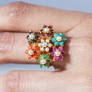 May include: A gold ring featuring a cluster of colorful flower designs. Each flower has a central pearl surrounded by petals in various colors, including blue, orange, green, pink, and turquoise. The ring is set on a hand.