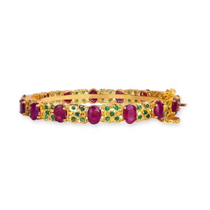 Könnte beinhalten: Ein goldenes Armreif-Armband mit roten und grünen Edelsteinen. Das Armband ist mit einem floralen Muster verziert.