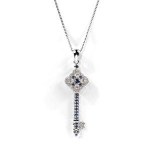 May include: A silver key pendant necklace featuring a clear and blue crystal design. The key has a square head with a floral pattern and a long, thin shaft.