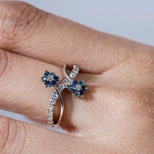 Könnte beinhalten: Ein silberner Ring mit zwei blauen Saphir-Blütenmotiven, die in einem zarten, gedrehten Band gefasst sind.