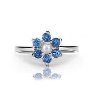 Può includere: Un anello d'argento con una perla bianca al centro e sei pietre preziose blu disposte a forma di fiore.