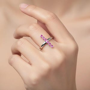 May include: A silver ring featuring a cross design. The cross is adorned with a series of pink gemstones. The ring is worn on a finger, showcasing its elegant and modern style. The gemstones are a vibrant pink color.