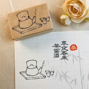 Puede incluir: Un sello de madera con un dibujo lineal negro de una tetera sobre una bandeja y dos tazas. Junto a él, un papel con un dibujo similar y texto japonés. Una flor de color crema en el fondo.