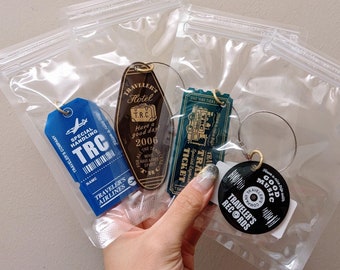 May include: Four keyrings in clear plastic bags. The keyrings feature different designs: a blue tag with "TRC" and "Traveler's Airlines", a brown hotel key, a green ticket, and a black record with "Good Music" and "Traveler's Records".