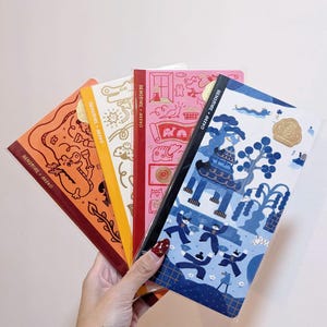 Peut inclure: Plusieurs cahiers colorés avec des couvertures originales. Les cahiers présentent des illustrations en orange, jaune, rose et bleu. Les couvertures affichent divers motifs, dont un animal de dessin animé, un bâtiment et d'autres éléments décoratifs. Le texte "BENSTORE + REKYO" est imprimé sur la tranche.