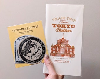 May include: A Traveler's Factory letterpress sticker set featuring a circular train graphic and a rectangular card with the text Train Trip From Tokyo Station and an illustration of the Tokyo Station Marunouchi Building in a clear plastic sleeve.