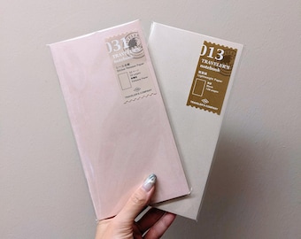 May include: Two Traveler's Company notebooks. One is a pale pink color, and the other is a light beige. Both notebooks have a rectangular shape and are sealed in clear plastic. The notebooks have a stamp-like label with Japanese text.