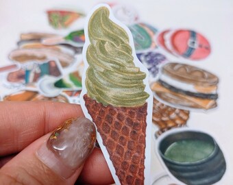 May include: A sticker of a matcha green tea ice cream cone with a brown waffle cone. The sticker is held by a hand, with other food-themed stickers in the background.