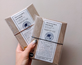 May include: Two passport-size Traveler's notebooks in brown paper packaging. Each notebook is wrapped in clear plastic and secured with a black or brown elastic band. The labels read "TRAVELER'S notebook PASSPORT SIZE".