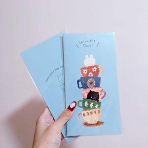 Puede incluir: Dos cuadernos azul claro con una ilustración caprichosa de tazas apiladas, cada una adornada con patrones únicos y un pequeño animal. El texto "Leisurely Days!" y "turn your life in" están impresos en las portadas.