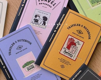 Op de afbeelding: Een verzameling reisnotitieboekjes in verschillende kleuren, waaronder roze, geel, lavendel en zwart. Elk notitieboekje heeft de tekst "TRAVELER'S NOTEBOOK" en unieke illustraties. De notitieboekjes liggen op een houten oppervlak.