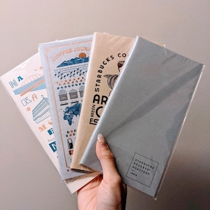 Op de afbeelding: Een hand houdt een stapel Starbucks-notitieboekjes vast. De notitieboekjes hebben verschillende ontwerpen, waaronder een brug, een berglandschap en het Starbucks Reserve Roastery-logo. De kleuren zijn voornamelijk blauw, beige en grijs.