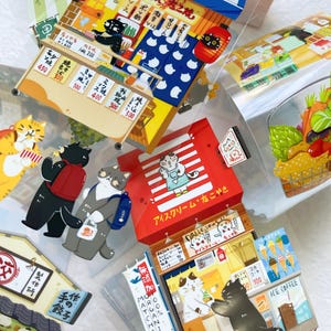 May include: A collection of colorful, illustrated stickers featuring cartoon cats in various settings. The stickers depict Japanese food stalls, ice cream shops, and other scenes with Japanese text and prices.