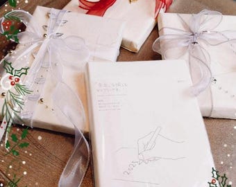 May include: Several wrapped gifts with ribbons and bows on a wooden tray. The gifts are wrapped in white paper and adorned with red, silver, and white ribbons. One gift has a red bow with a gold leaf accent. A white notebook with the year 2025 is visible.