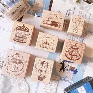 Puede incluir: Una colección de sellos de madera con ilustraciones caprichosas. Los diseños incluyen un carrusel, una carpa de circo, un tren, un globo aerostático y un personaje con globos. Los sellos están hechos de madera de color claro y presentan dibujos grabados detallados.