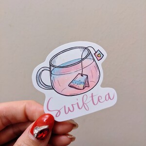 May include: A sticker featuring a clear teacup with pink and blue liquid and a tea bag. The word "Swifttea" is written in pink cursive below the cup. The sticker has a white border.