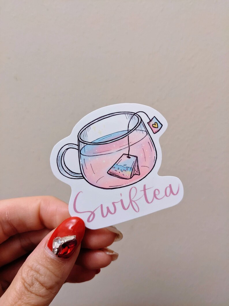 May include: A sticker featuring a clear teacup with pink and blue liquid and a tea bag. The word "Swifttea" is written in pink cursive below the cup. The sticker has a white border.