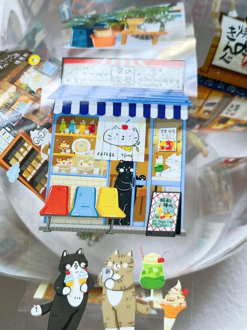 May include: A colorful sticker sheet featuring a cat-themed coffee shop scene. The shop has a blue awning, a sign that says "COFFEE TIME", and colorful drinks and pastries. There are also cat characters holding drinks and taking photos.