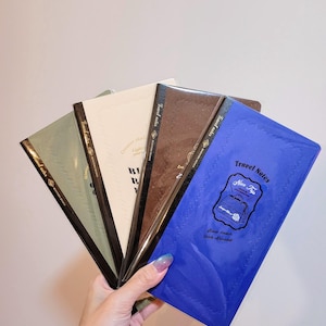 Könnte beinhalten: Eine Hand hält einen Fächer aus Reisetagebüchern in verschiedenen Farben: Grün, Creme, Braun und Blau. Das blaue Notizbuch trägt den Text "Travel Notes" und "Nice Trip". Die Notizbücher haben schwarze Rücken.