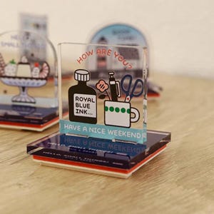 May include: Clear acrylic stamp with a cartoon design. The stamp features a black ink bottle, a pen, scissors, and a mug with green dots. Text includes "HOW ARE YOU?" and "HAVE A NICE WEEKEND".