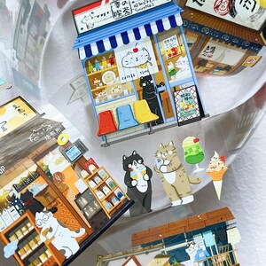 May include: A collection of colorful cat-themed stickers featuring Japanese-style shops and cafes. The stickers include illustrations of cats, food, and Japanese text. The stickers are in various shapes and sizes.