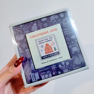 May include: A boxed calendar for 2026 with a winter theme. The calendar features a stamp-like design with a knitted hat and Christmas tree illustration. The box has a purple background with winter-themed illustrations and the text "Enjoy the small things."