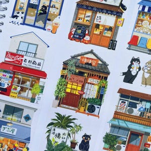 May include: A spiral-bound notebook featuring colorful sticker illustrations of Japanese storefronts and shops. The stickers depict various businesses with detailed facades, including restaurants and shops, with Japanese text and cartoon characters.