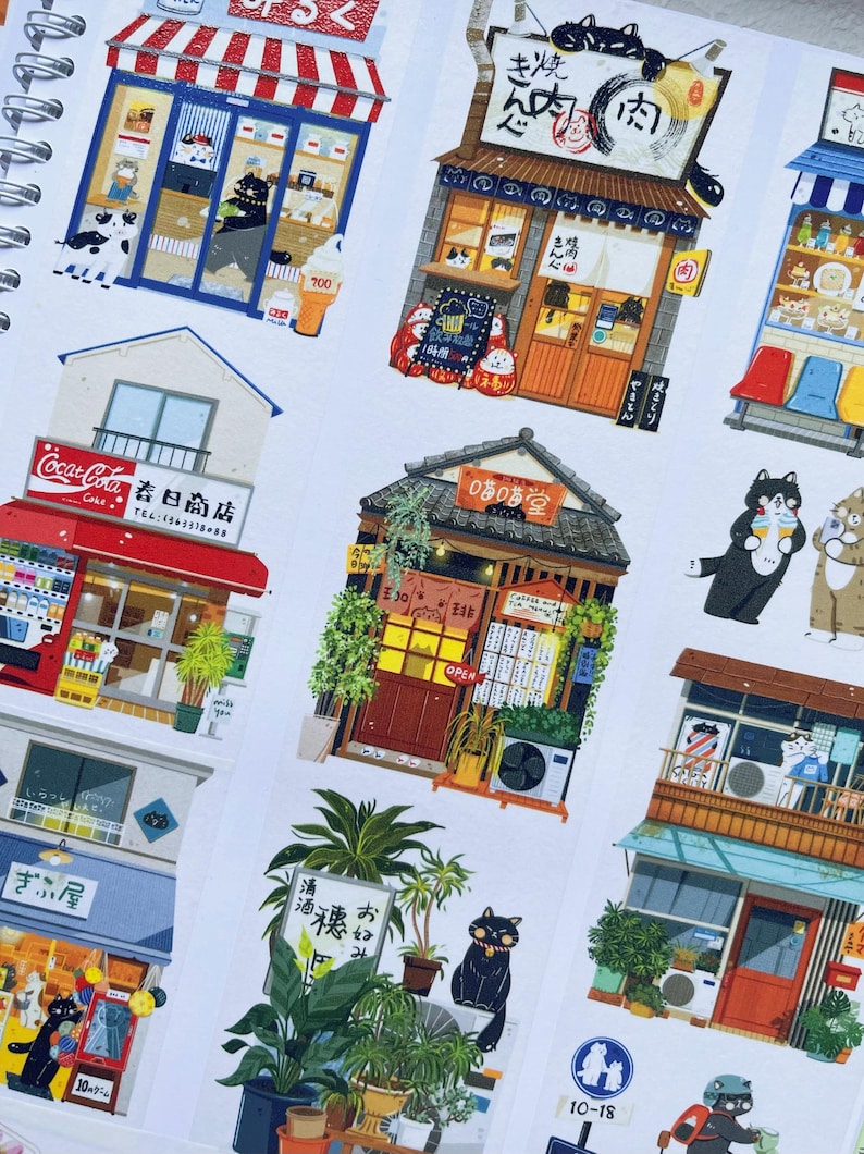 May include: A spiral-bound notebook featuring colorful sticker illustrations of Japanese storefronts and shops. The stickers depict various businesses with detailed facades, including restaurants and shops, with Japanese text and cartoon characters.
