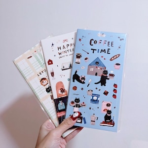Puede incluir: Tres cuadernos ilustrados con diferentes temas. Un cuaderno tiene el texto "Coffee Time" sobre un fondo azul con ilustraciones de gatos. Otro dice "Happy Winter" con ilustraciones con temática invernal. El tercer cuaderno tiene el texto "Let's" con ilustraciones.