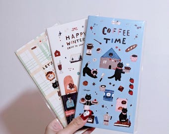 Puede incluir: Tres cuadernos ilustrados con diferentes temas. Un cuaderno tiene el texto "Coffee Time" sobre un fondo azul con ilustraciones de gatos. Otro dice "Happy Winter" con ilustraciones con temática invernal. El tercer cuaderno tiene el texto "Let's" con ilustraciones.
