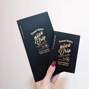 Könnte beinhalten: Zwei schwarze Reisetagebücher mit goldfarbenen Buchstaben. Das größere Notizbuch steht aufrecht, das kleinere wird in einer Hand gehalten. Der Text auf den Notizbüchern lautet "Travel Notes nice Trip" und "SLOW DOWN LOOK AROUND".