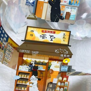 May include: A whimsical, layered papercraft scene featuring a coffee shop and an ice cream stand, both with cat characters. The coffee shop has shelves of products and a menu. The ice cream stand offers ice cream cones. Japanese text is visible.