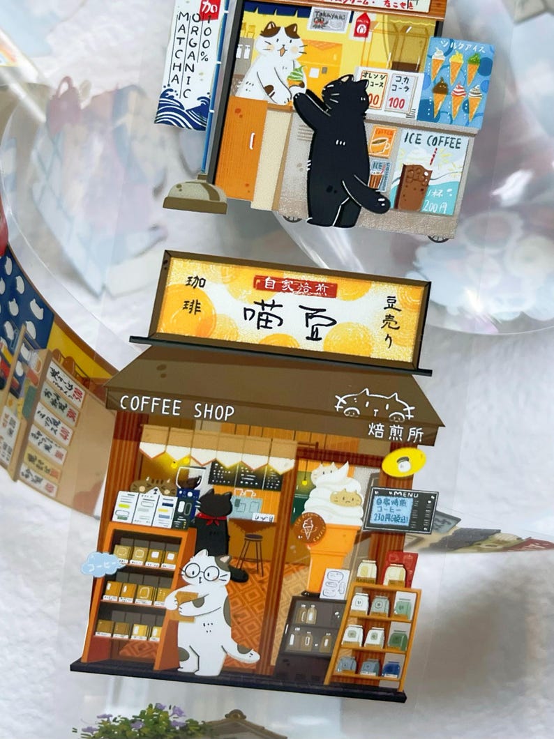 May include: A whimsical, layered papercraft scene featuring a coffee shop and an ice cream stand, both with cat characters. The coffee shop has shelves of products and a menu. The ice cream stand offers ice cream cones. Japanese text is visible.