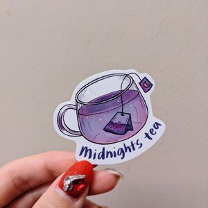 May include: A sticker featuring a clear glass teacup filled with purple liquid and a tea bag. The cup has a handle and the words "Midnights tea" in a curved font. A crescent moon is on the tea bag tag.