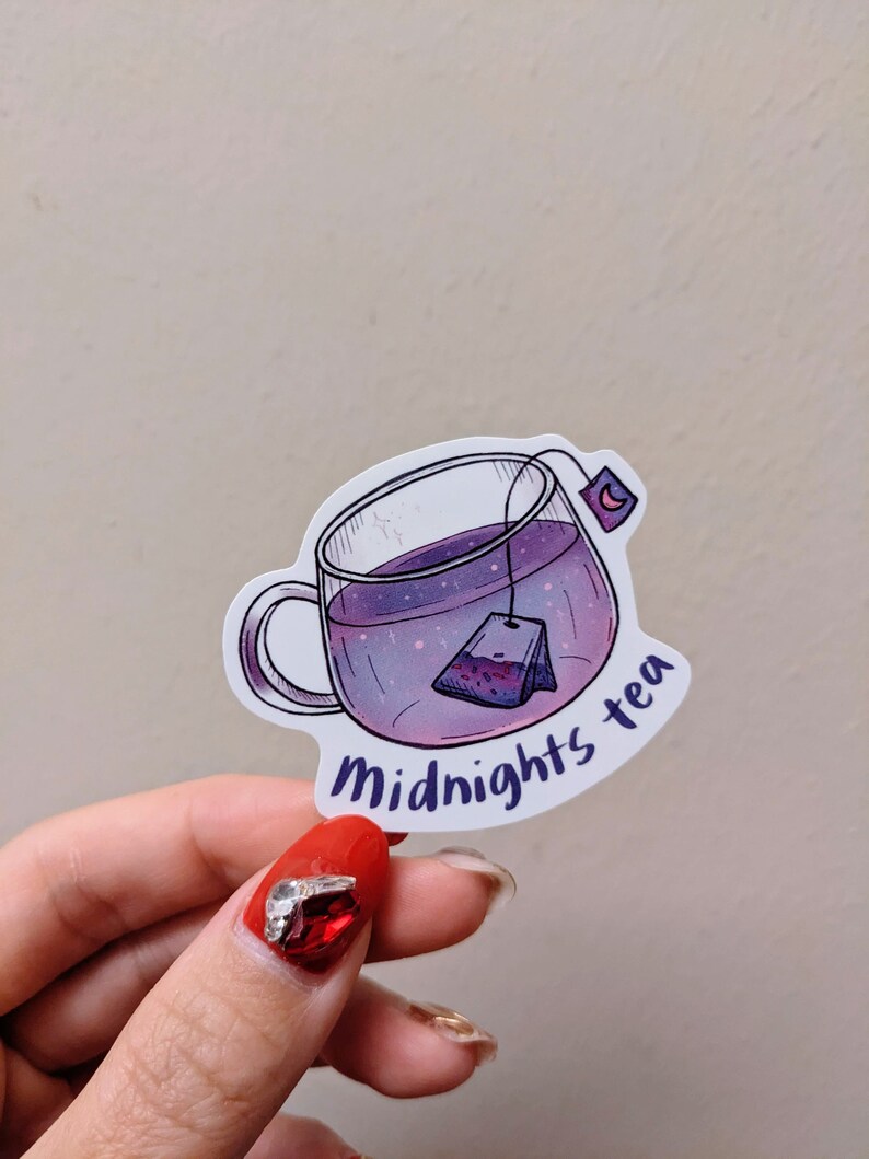 May include: A sticker featuring a clear glass teacup filled with purple liquid and a tea bag. The cup has a handle and the words "Midnights tea" in a curved font. A crescent moon is on the tea bag tag.