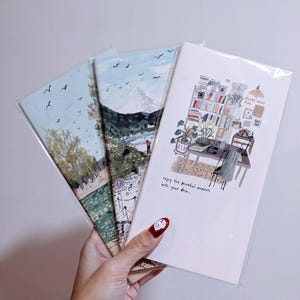 Pode incluir: Três cadernos ilustrados com paisagens, cada um em uma capa de plástico. Os desenhos retratam uma cena de montanha, uma vista à beira-mar e um interior. O texto diz: "enjoy the peaceful moments with your desk..."