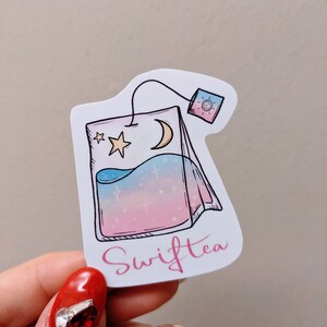 May include: A sticker in the shape of a tea bag with a celestial design. The tea bag features a crescent moon, stars, and a gradient of blue, pink, and white. The word "Swifttea" is written in pink script.