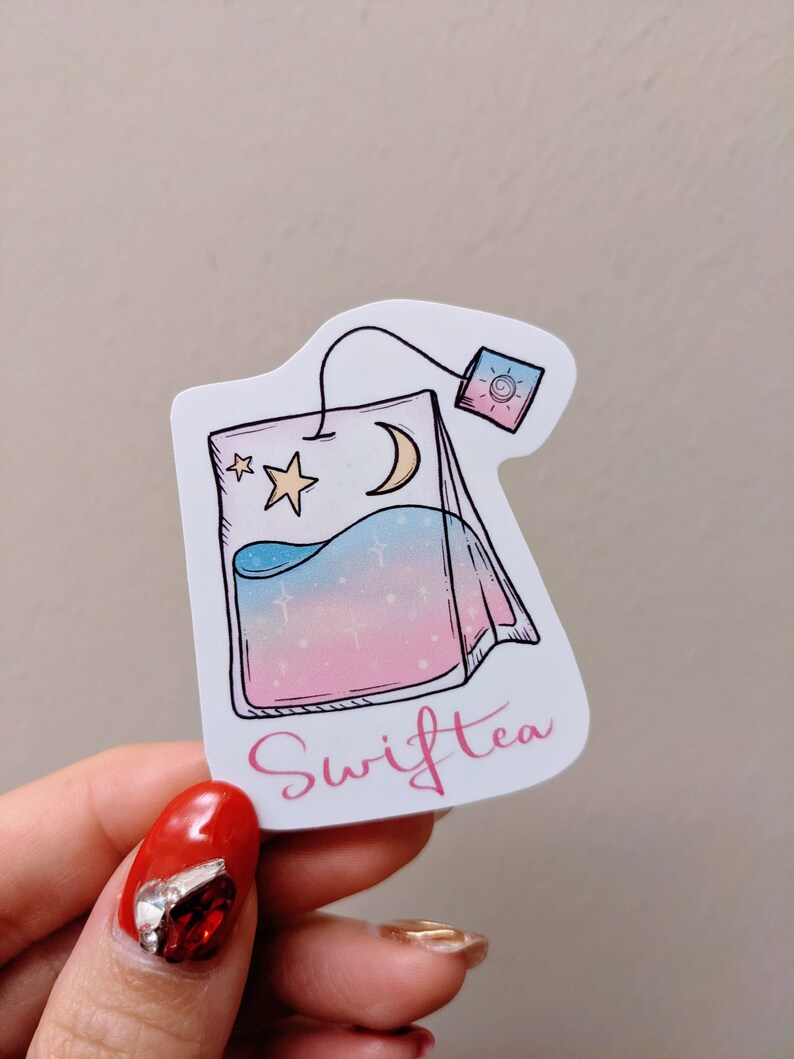 May include: A sticker in the shape of a tea bag with a celestial design. The tea bag features a crescent moon, stars, and a gradient of blue, pink, and white. The word "Swifttea" is written in pink script.