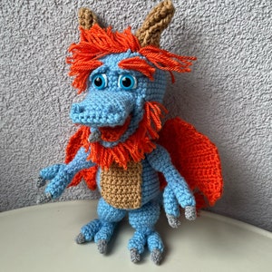 Op de afbeelding: Een handgemaakt gehaakt draakspeelgoed in tinten blauw, oranje, bruin en grijs. De draak heeft grote blauwe ogen, oranje haar en vleugels. De draak staat op een wit oppervlak.