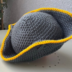 May include: A gray crocheted hat with a yellow trim. The hat is shaped like a pirate hat.