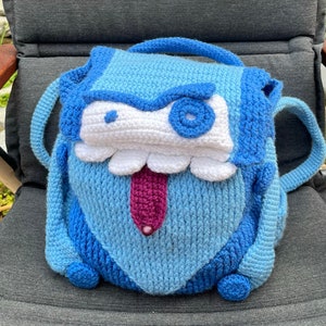 May include: A blue crocheted backpack shaped like a monster with a white mouth, a purple tongue, and a blue eye. The backpack has two straps.
