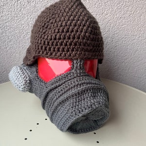 May include: A handmade crochet hat and scarf set in shades of brown and gray. The hat has a pointed top and ear flaps, while the scarf is a matching gray. The set is designed to fit a small object, possibly a mug or a small head.